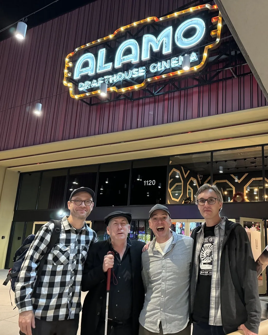 Erik Horn, Jim Knipfel in a flat cap holding his white cane, Brandon Potter and Brad Parrett, smiling under the neon sign of an Alamo Drafthouse Cinema at night